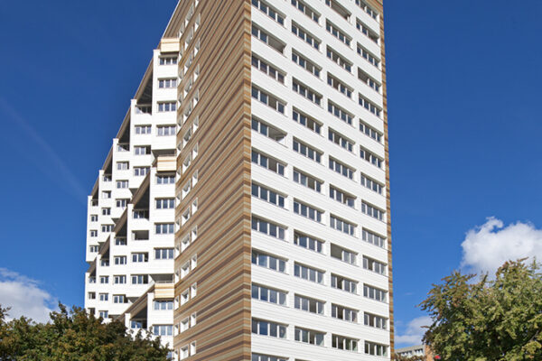 Hochhaus mit modernem Design, umgeben von Bäumen und blauem Himmel, im Vordergrund ein gepflasterter Platz.
