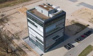 Moderne Bürogebäude mit Glasfassade, umgeben von unbebautem Gelände und parkenden Autos.