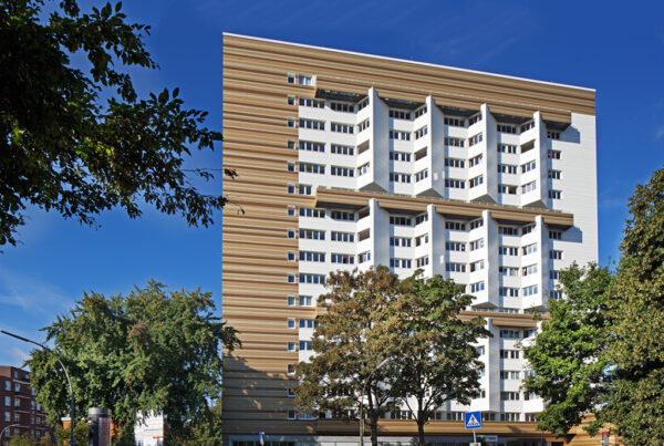 Modernes Wohngebäude mit auffälliger Fassade, umgeben von Bäumen und blauem Himmel. Parkplatz im Vordergrund.
