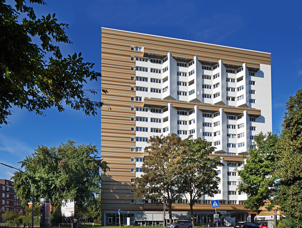 Modernes Wohngebäude mit auffälliger Fassade, umgeben von Bäumen und blauem Himmel. Parkplatz im Vordergrund.