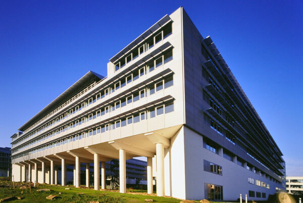 Modernes Bürogebäude mit mehreren Etagen, großen Fenstern und Säulen, vor blauem Himmel.