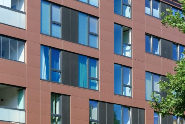 Moderne Wohnhausfassade mit großen Fenstern, braunen Fliesen und einem begrünten Bereich im Vordergrund.