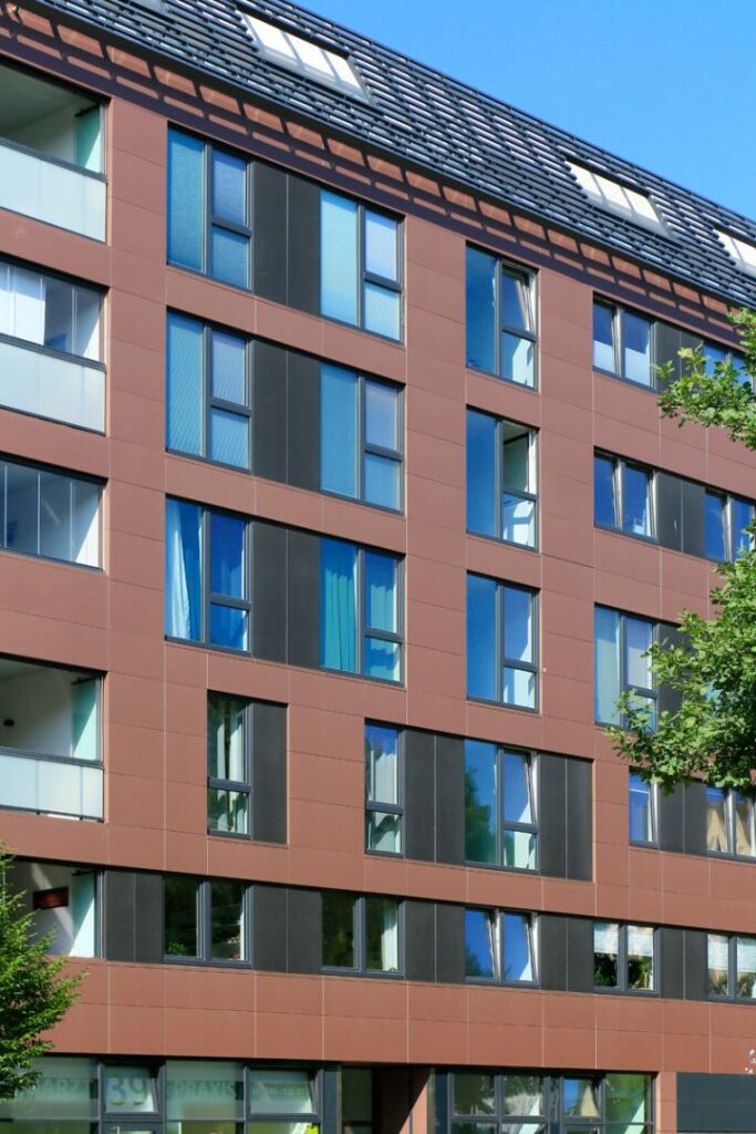 Moderne Wohnhausfassade mit großen Fenstern, braunen Fliesen und einem begrünten Bereich im Vordergrund.