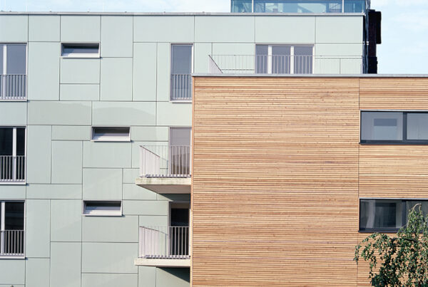 Moderne Wohnanlage mit Holz- und Glasfassade, begrüntem Außenbereich und Balkonen. Heller Himmel im Hintergrund.