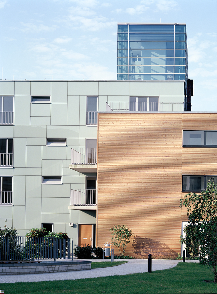 Moderne Wohnanlage mit Holz- und Glasfassade, begrüntem Außenbereich und Balkonen. Heller Himmel im Hintergrund.
