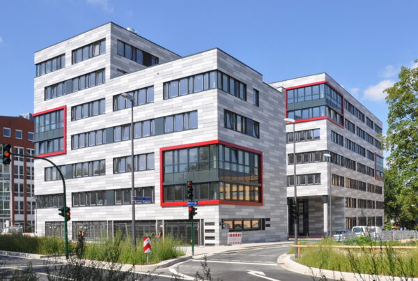 Modernes Bürogebäude mit grauer Fassade und roten Akzenten, umgeben von Grünflächen und Verkehrssignalen.