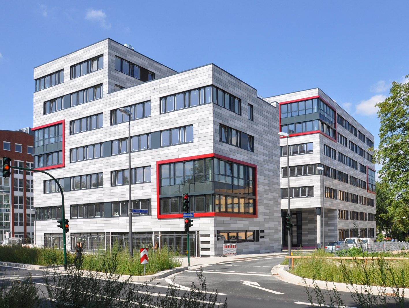 Modernes Bürogebäude mit grauer Fassade und roten Akzenten, umgeben von Grünflächen und Verkehrssignalen.