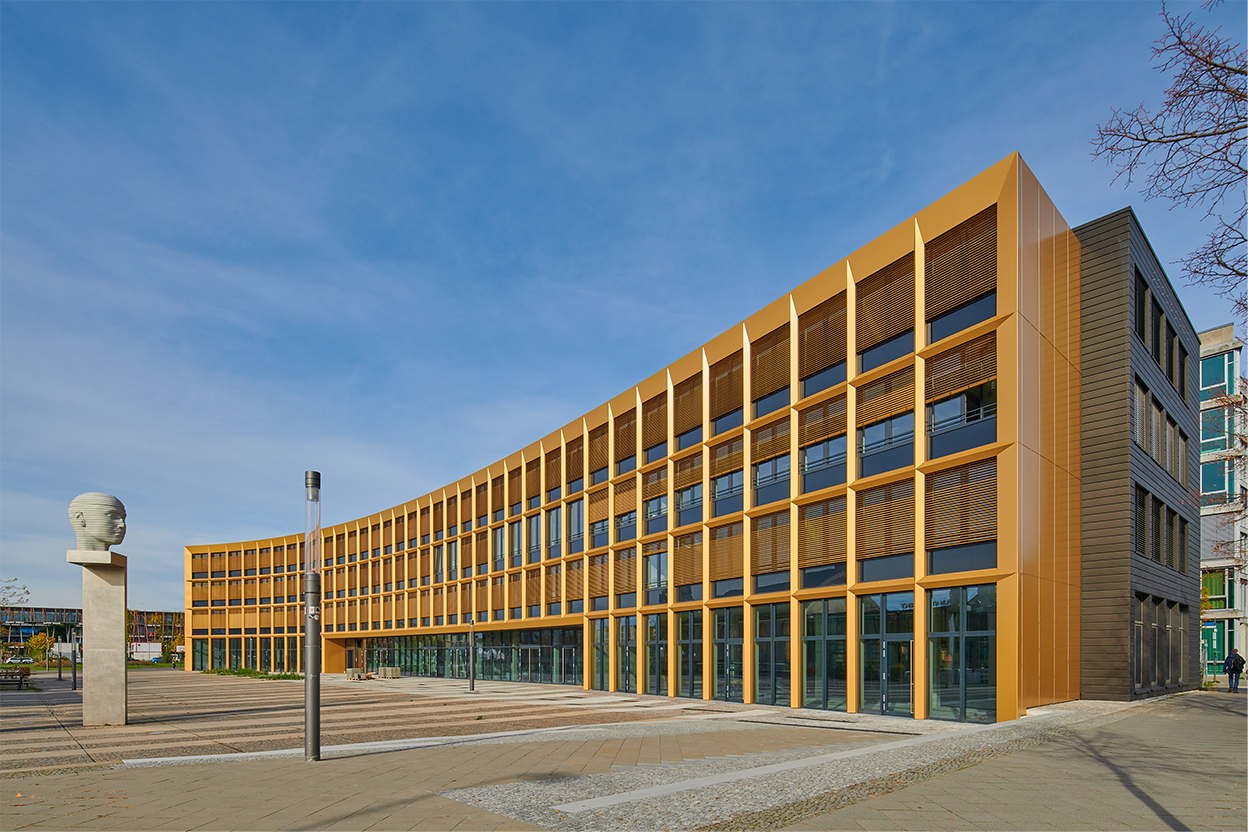 Moderne Architektur eines Gebäudes mit großer Fensterfront und einer Skulptur im Vordergrund, unter blauem Himmel.