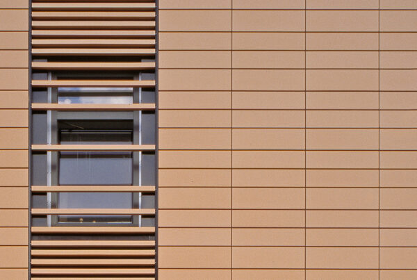 Fenster mit horizontalen Lamellen in einer beige-farbenen Wandverkleidung aus rechteckigen Platten.