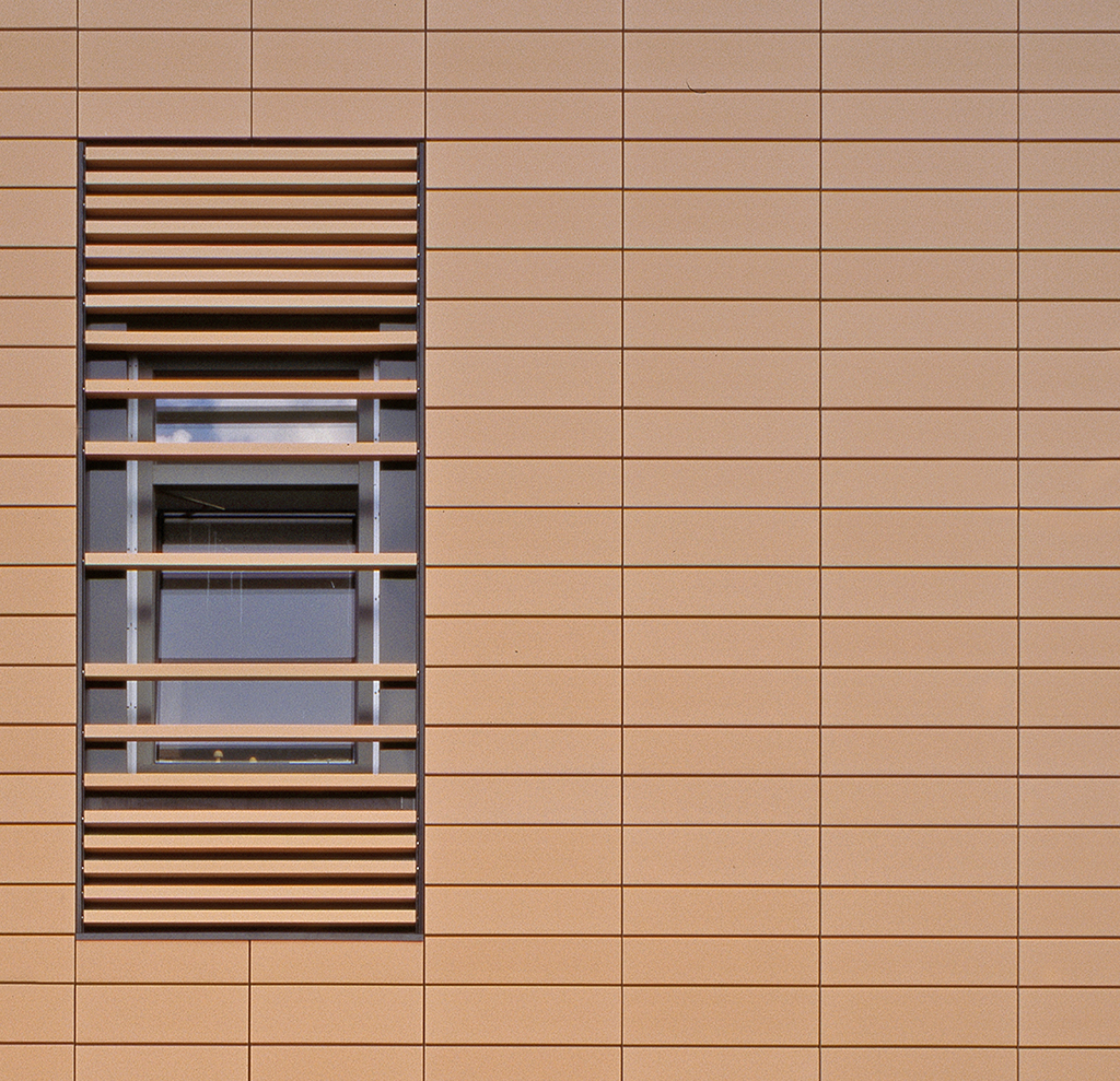 Fenster mit horizontalen Lamellen in einer beige-farbenen Wandverkleidung aus rechteckigen Platten.