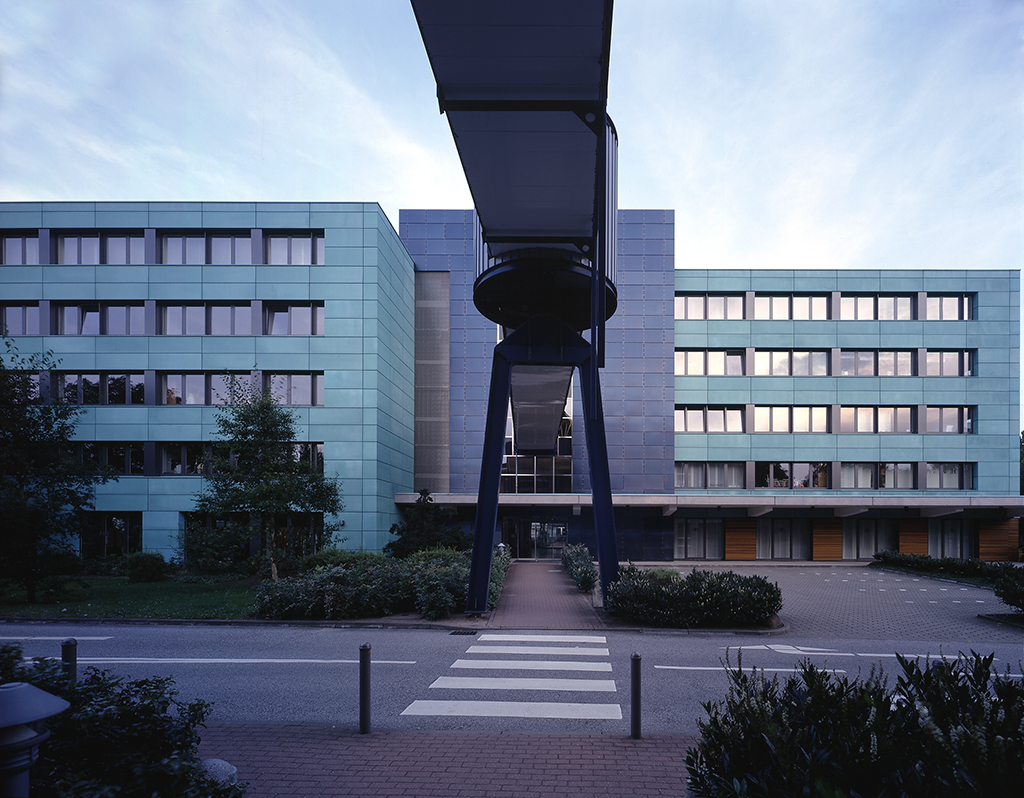 Moderne Gebäude mit Glasfassade und überdachtem Gehweg, umgeben von Bäumen und Grünflächen.