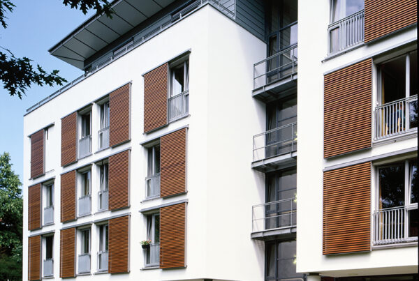 Moderne Wohngebäude mit weißen Wänden, Holzjalousien und Balkonen, umgeben von Rasen und Bäumen.
