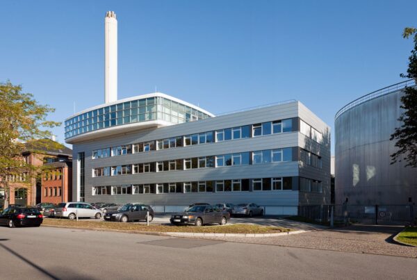 Moderne Bürogebäude mit Glasfassade und Schornstein, umgeben von parkenden Autos und Bäumen. Klarer blauer Himmel.