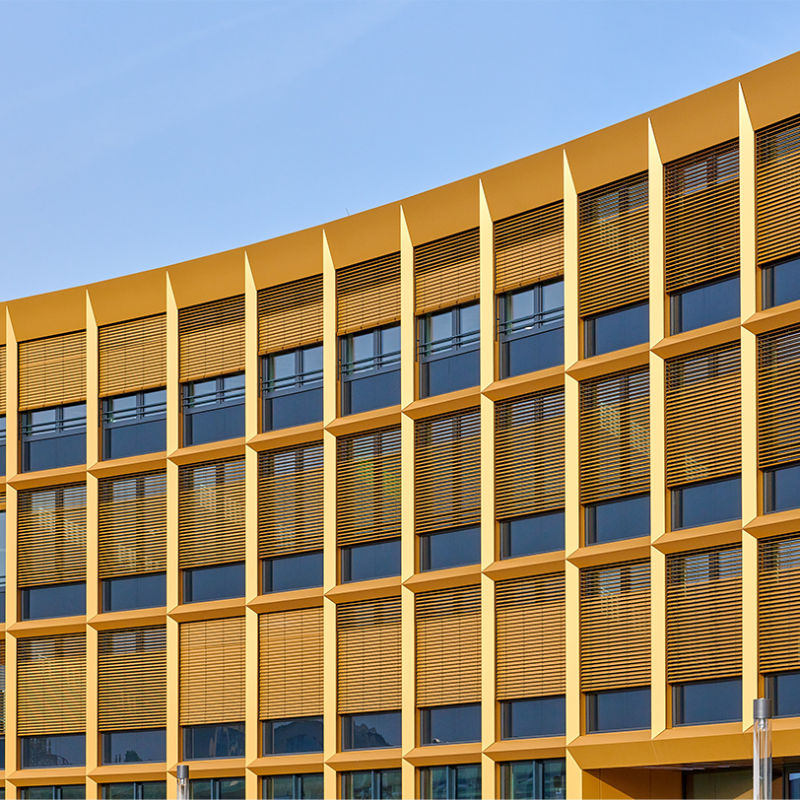 Moderne Gebäudeansicht mit gelben Fassadenelementen und horizontalen Jalousien vor großen Fenstern.