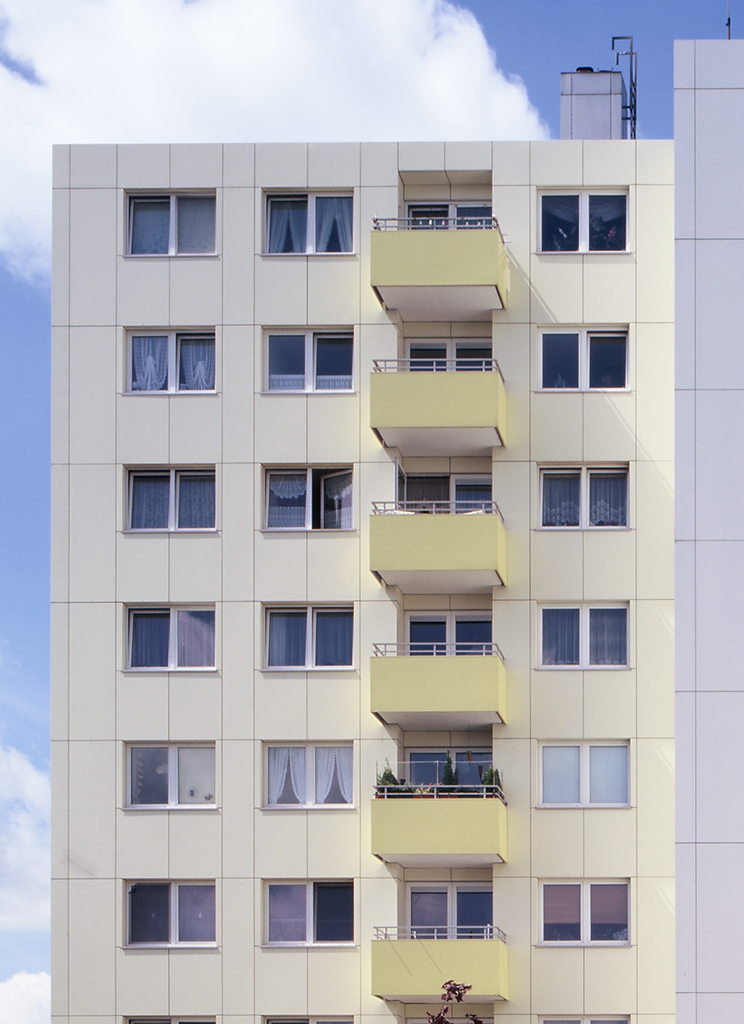 Helles Mehrfamilienhaus mit gelben Balkonen und mehreren Fenstern, vor einem blauen Himmel mit Wolken.