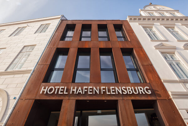 Moderne Fassade des Hotel Hafen Flensburg mit rostigem Metall und angrenzendem klassischem Gebäude.