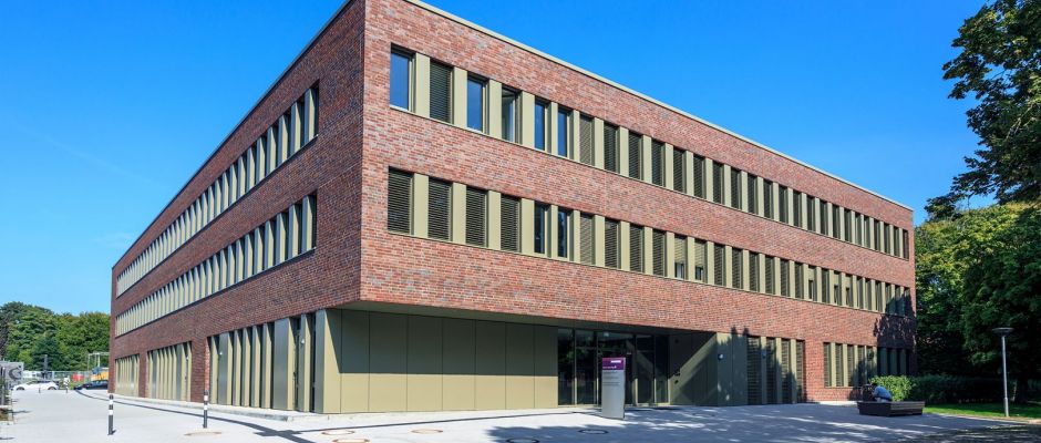 Modernes, dreigeschossiges Gebäude mit roten Ziegeln und großen Fenstern, umgeben von Grünflächen und klarem Himmel.