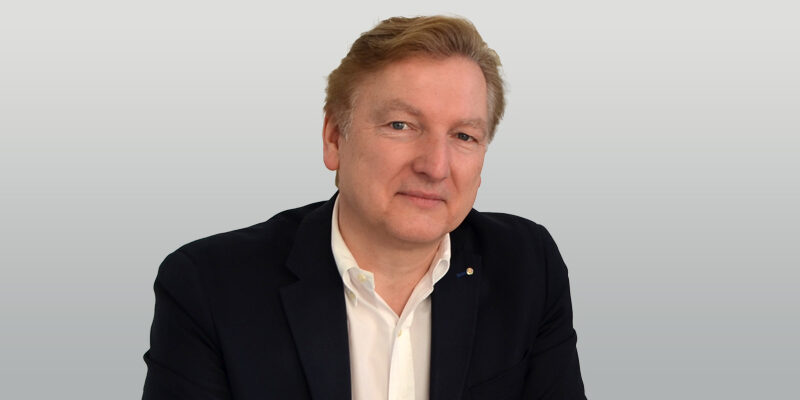 Portrait eines Mannes mit blonden Haaren, der in einem schwarzen Blazer und weißen Hemd lächelt, vor einem grauen Hintergrund.