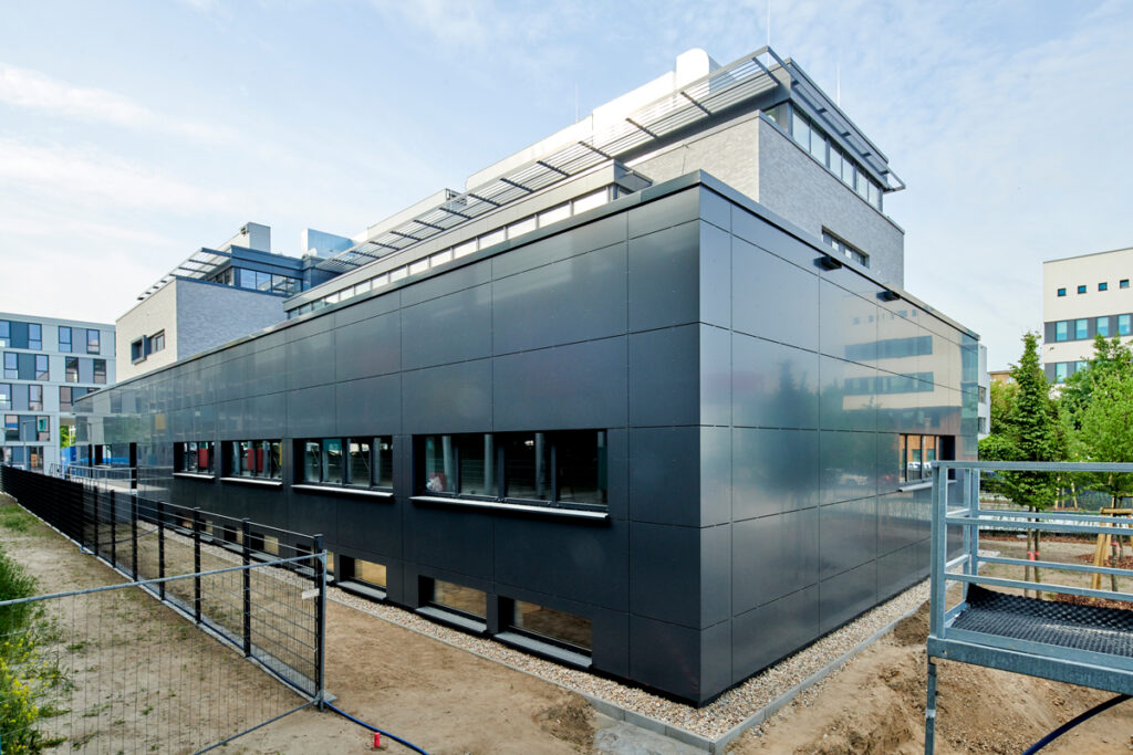 Modernes Gebäude mit schwarzer Fassade, großen Fenstern und umliegendem Bauzaun. Sonnige Tagesaufnahme.