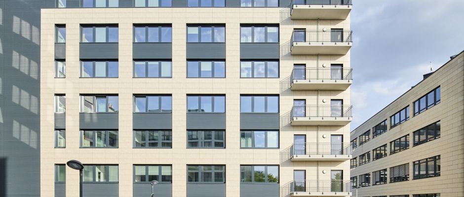 Moderne Wohngebäude mit großen Fenstern und Balkonen, umgeben von weiteren Gebäuden und einem klaren Himmel.