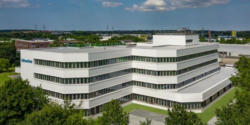 Moderne Bürogebäude mit mehreren Etagen und großen Fenstern, umgeben von Grünflächen und Industrieanlagen.