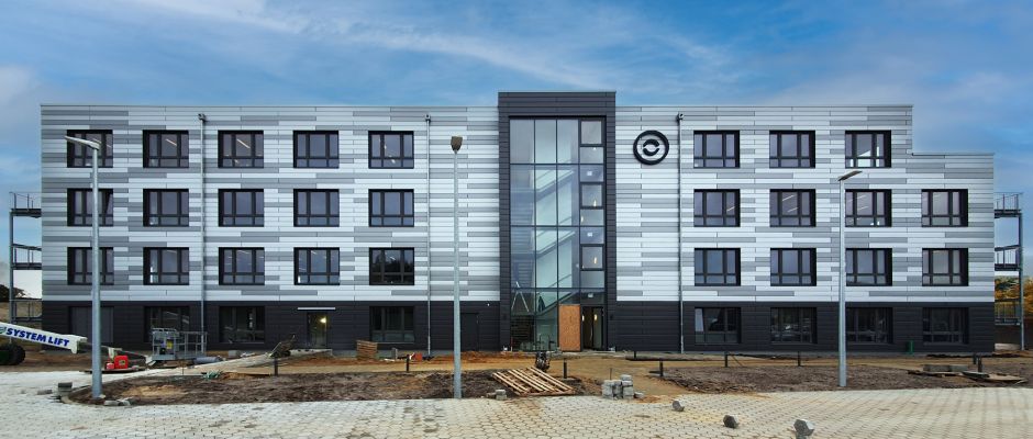 Neues, modernes Gebäude mit mehreren Fenstern und einer gläsernen Eingangsfront, umgeben von Baustellenarbeiten.
