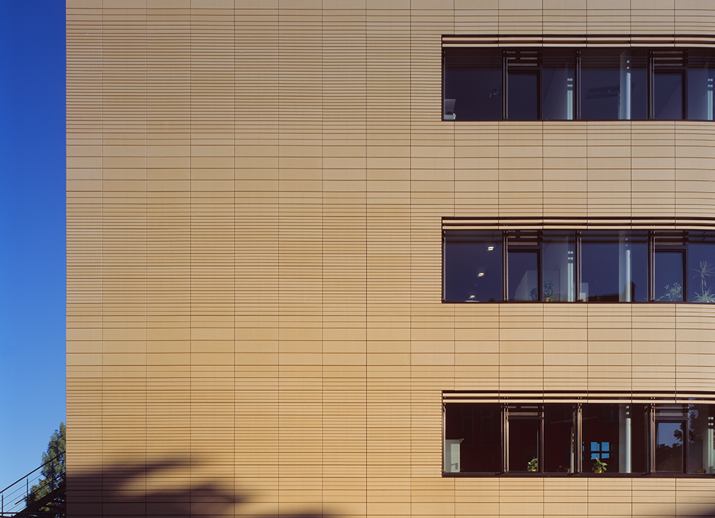 Fassade eines modernen Gebäudes mit horizontalen Linien und großen Fenstern, blauer Himmel im Hintergrund.