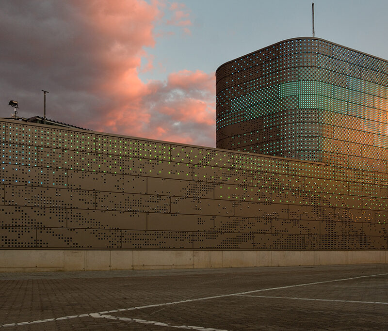 Moderne Architektur mit einer auffälligen Fassade aus perforiertem Metall und bunten Lichtakzenten vor einem bewölkten Himmel.