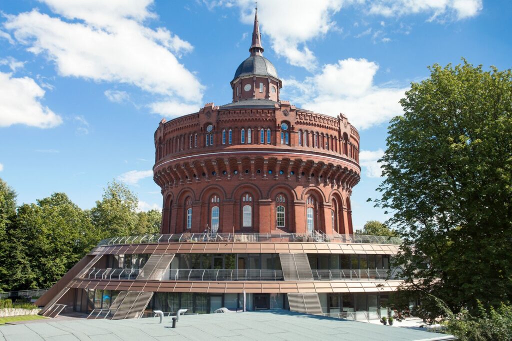 Runder, roter Backsteinbau mit spitzem Dach und moderner Glasarchitektur, umgeben von Bäumen und blauem Himmel.