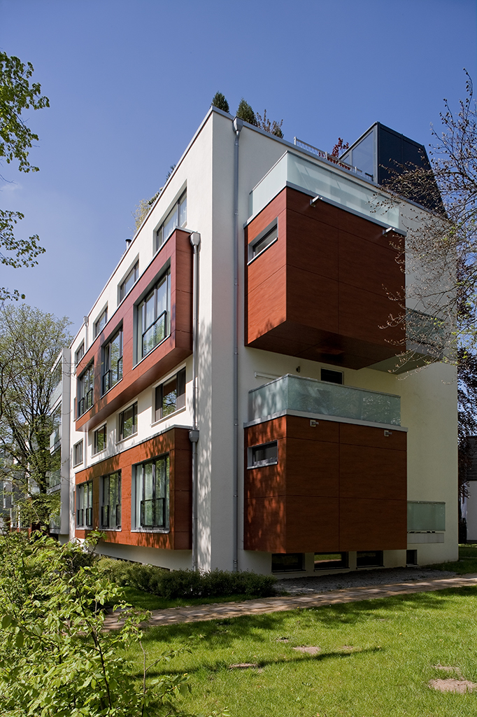 Modernes Wohngebäude mit Holzverkleidung und großen Fenstern in einer grünen Umgebung unter blauem Himmel.