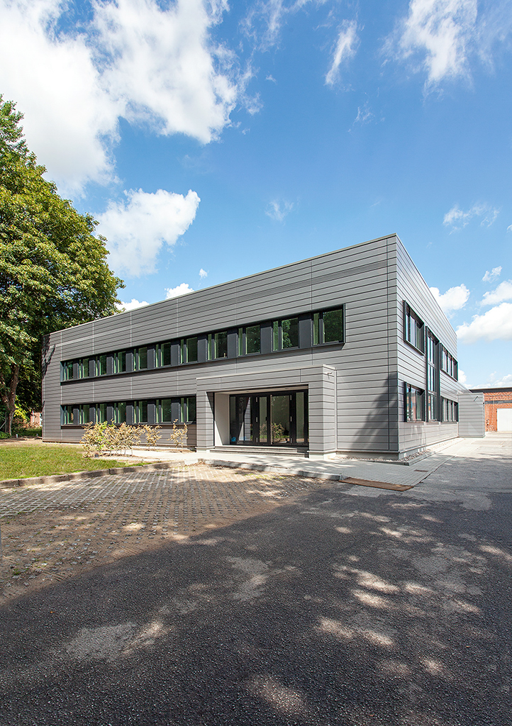 Modernes, zweigeschossiges Gebäude mit großen Fenstern, umgeben von Grünflächen und blauem Himmel.