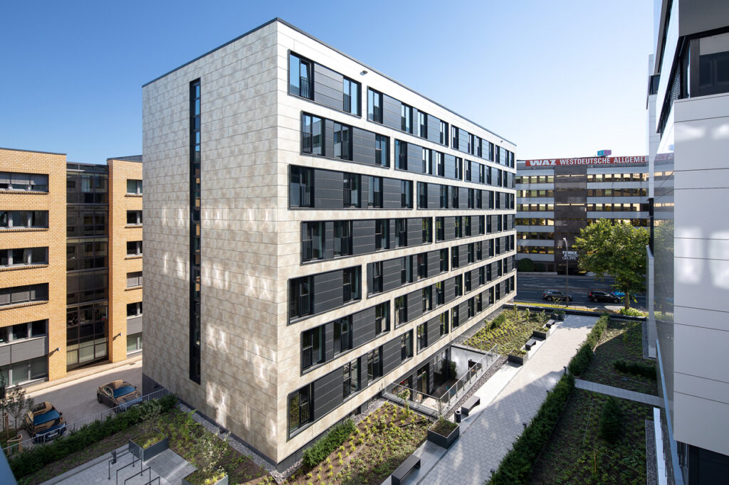 Moderne Wohngebäude mit Glasfenstern und begrünten Außenbereichen, vor blauem Himmel und urbanem Hintergrund.