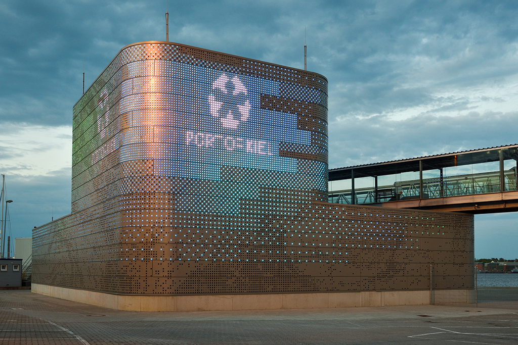 Modernes Gebäude mit einer perforierten Metallfassade und einem digitalen Recycling-Symbol, unter bewölktem Himmel.