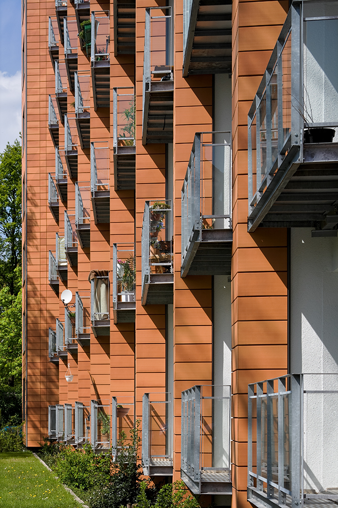Fassade eines modernen Wohngebäudes mit orangefarbenen Paneelen und mehreren Balkonen, umgeben von Grünflächen.