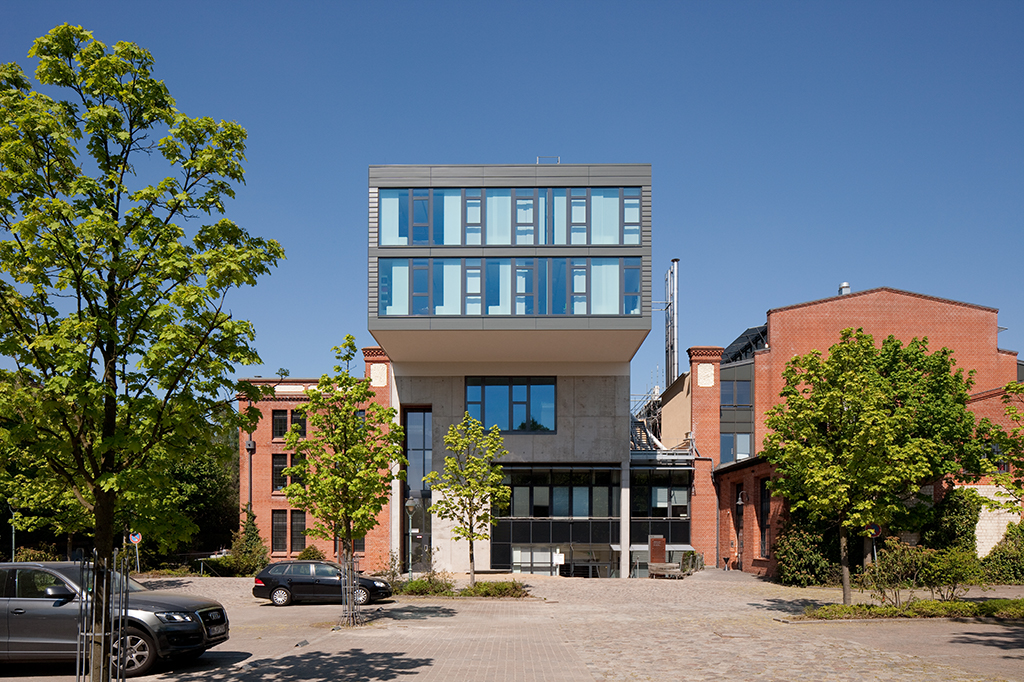 Modernes Gebäude mit Glasfront, umgeben von Bäumen und historischen Backsteinhäusern, bei klarem blauen Himmel.