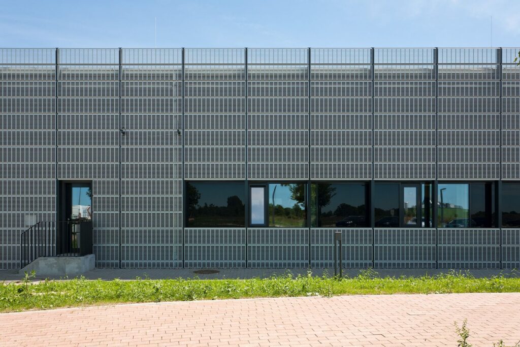 Fassade eines modernen Gebäudes mit Metallgitter und großen Fenstern, umgeben von Grünflächen und einem gepflasterten Weg.