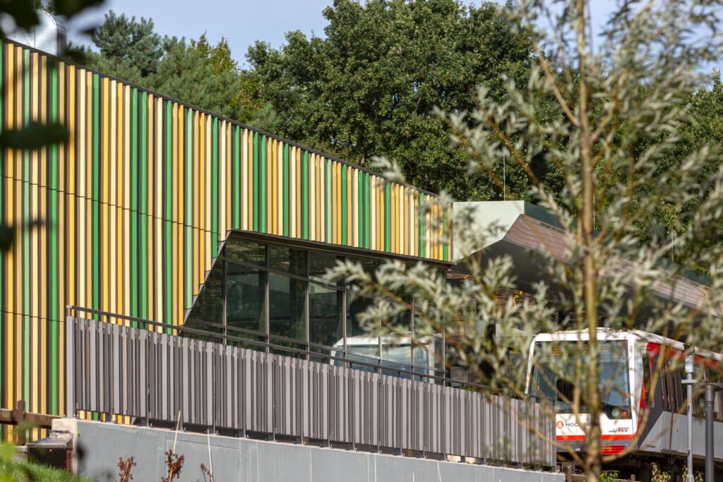 Moderne Haltestelle mit buntem, vertikal gestreiftem Fassadendesign und Zug auf der Plattform, umgeben von Bäumen.
