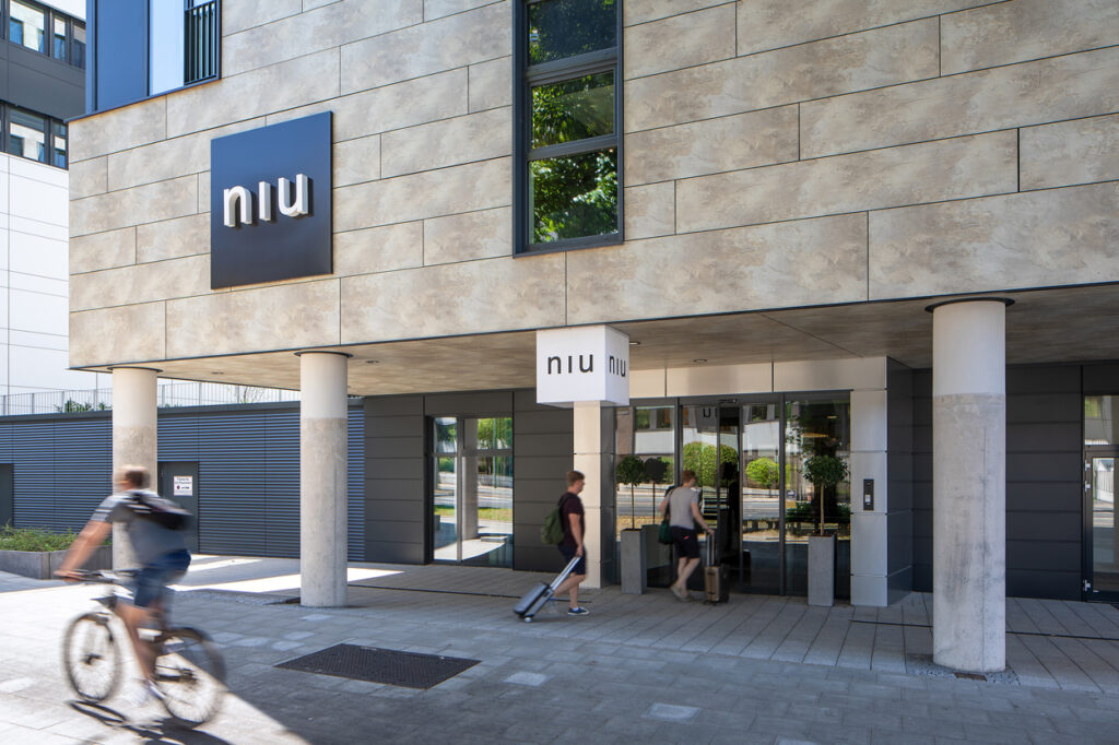 Eingang zum niu Hotel mit Gästen, die einchecken, und einem Radfahrer, der vorbeifährt. Modernes Architekturdesign.