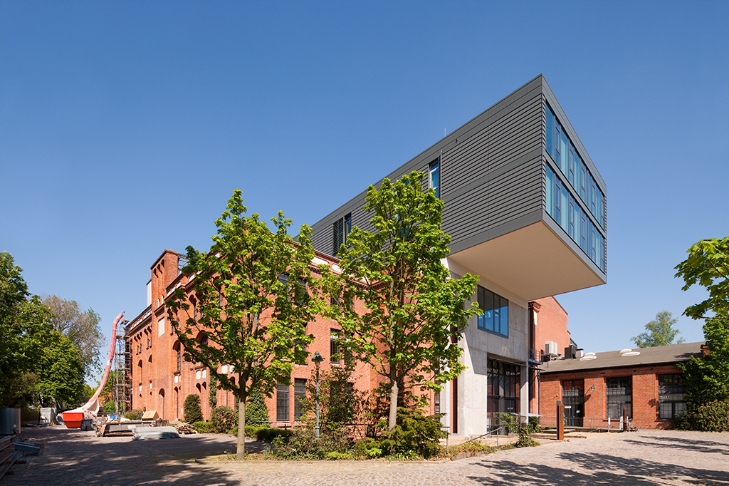 Moderne Gebäude mit überstehendem Obergeschoss, umgeben von Bäumen und historischen Ziegelmauern, blauer Himmel.