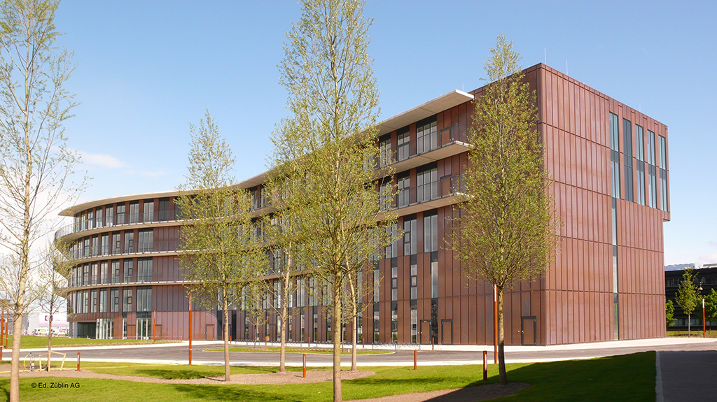 Moderne, mehrstöckige Bürogebäude mit großen Fenstern und umgebenden Bäumen, unter blauem Himmel.
