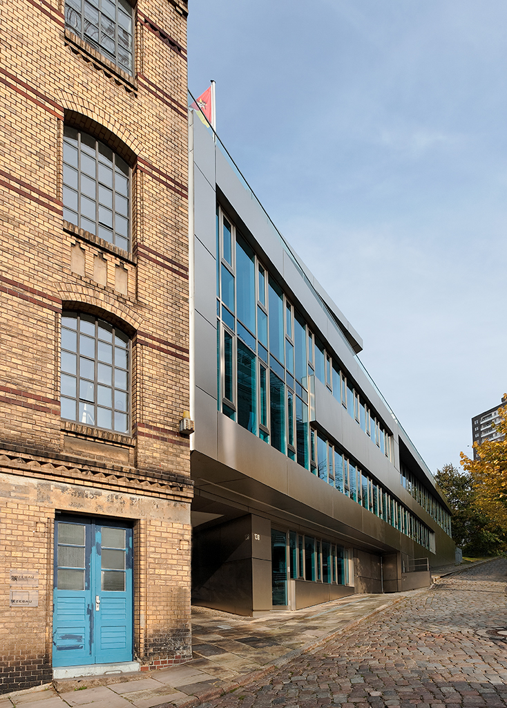 Modernes Gebäude mit Glasfassade neben einem historischen Backsteingebäude, gepflasterte Straße im Vordergrund.