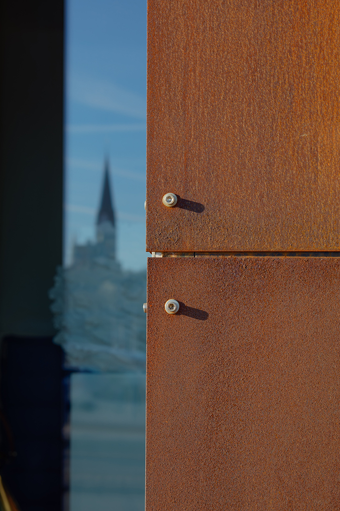 Detailaufnahme einer rostigen Metalloberfläche mit Schrauben, im Hintergrund ist ein Kirchturm zu sehen.
