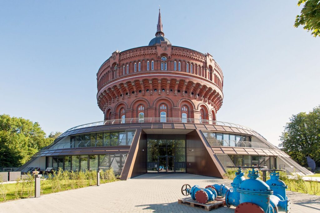 Ehemaliger Wasserturm mit modernen Anbauten, umgeben von grünen Flächen und blauen Wasserrohren im Vordergrund.