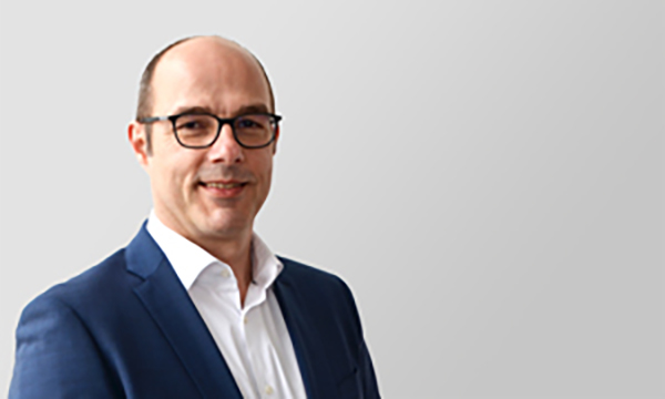 Mann mit Brille und Anzug lächelt vor einem hellgrauen Hintergrund.