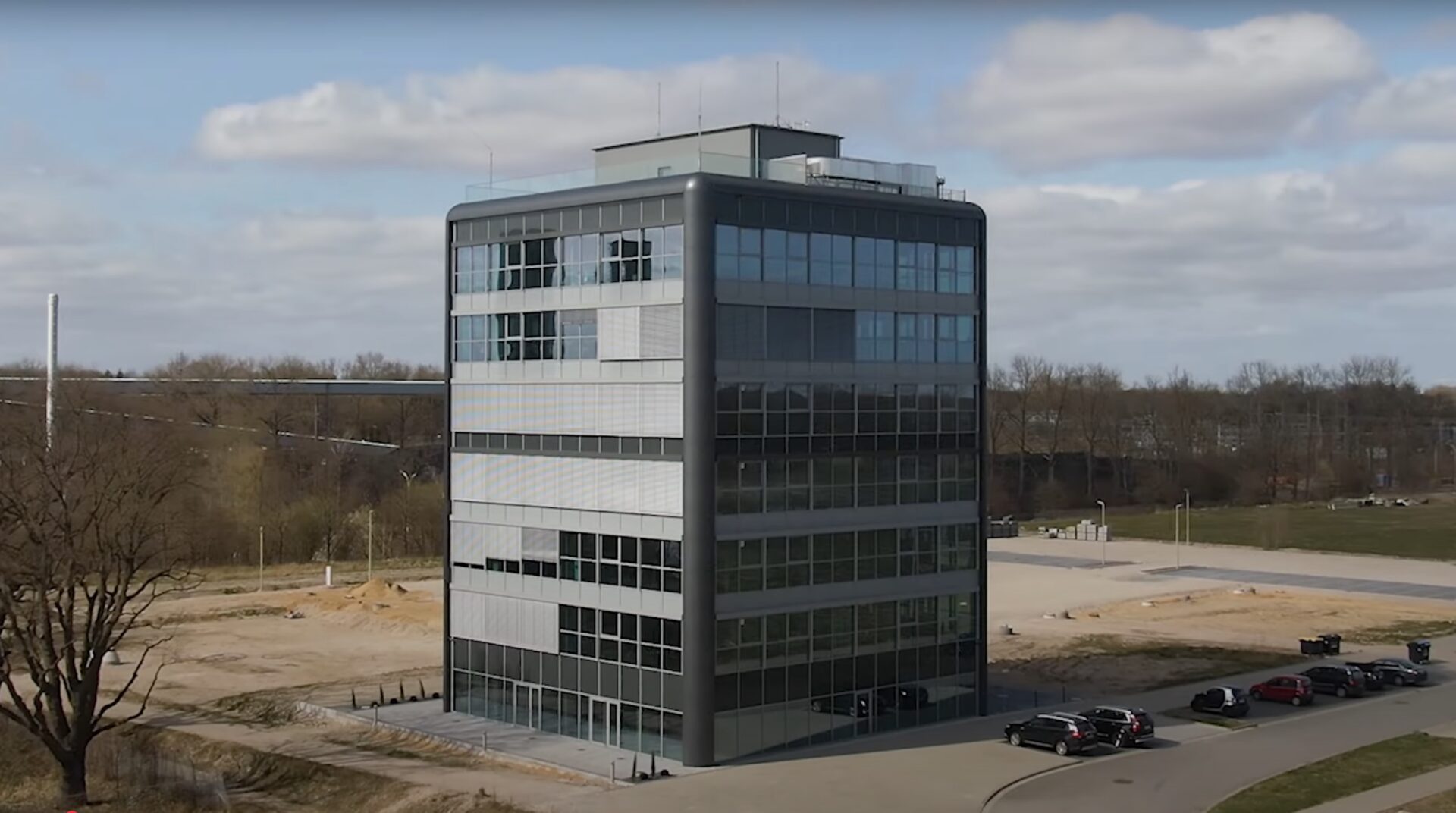 Modernes Bürogebäude mit Glasfassade, umgeben von Freiflächen und Parkplätzen, unter bewölktem Himmel.