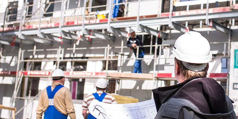 Bauarbeiter mit Helmen und Plänen stehen vor einem Gerüst und beobachten die Arbeiten an einem Gebäude.